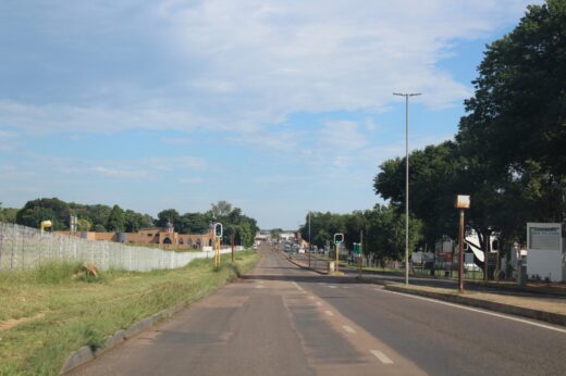 A quiet two-lane road extends into the distance, bordered by green grass and trees under a partly cloudy sky. Sparse traffic lights and buildings are visible.