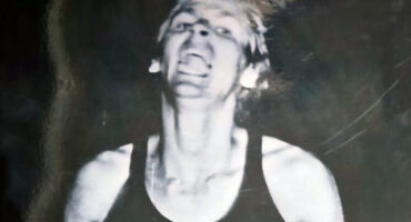 A black-and-white photo of a person with short hair and a tank top, captured mid-movement with a vivid expression, conveying intense energy.