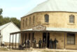 A group of people in Old West attire stand outside a rustic stone building labeled "J.W. Henwood Direct Importer," conveying a historical, frontier town feel.