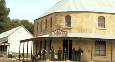A group of people in Old West attire stand outside a rustic stone building labeled "J.W. Henwood Direct Importer," conveying a historical, frontier town feel.