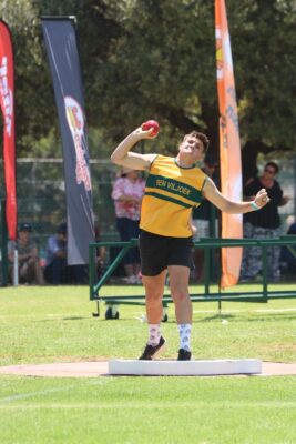 Athlete in yellow and green throwing a shot put in an outdoor stadium. Flags and spectators in the background; focus on determination and strength.