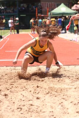 Athlete in yellow jersey lands in sand pit during a long jump, sand scattering. Her expression shows focus. Spectators and officials in the background.