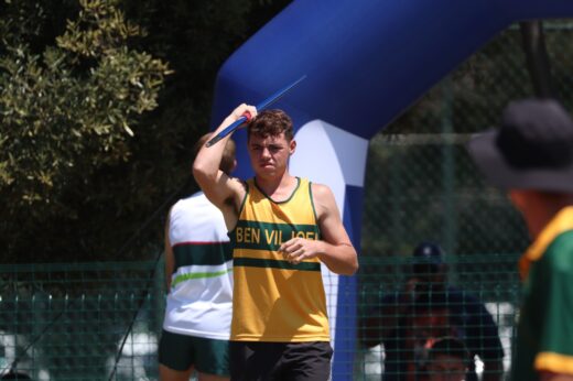 Athlete holding a javelin prepares to throw in bright sunlight, wearing a yellow and green tank top. The scene exudes focus and concentration.