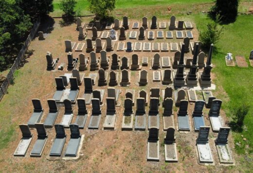 Aerial view of a cemetery with numerous rows of gravestones and tombs on a grassy field. The layout is orderly, evoking a solemn, peaceful atmosphere.