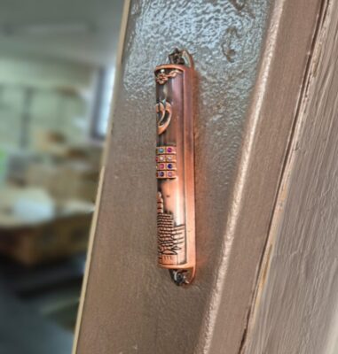 Bronze mezuzah affixed to a doorpost, adorned with intricate designs and colorful jewels. Background shows a softly blurred interior room.