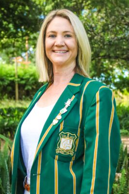 Smiling woman outdoors wearing a green and yellow striped blazer with a crest and pins, against a lush, leafy background. Bright, cheerful ambiance.