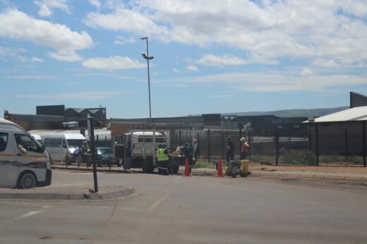 A street scene shows workers in safety vests and helmets managing traffic cones near parked vehicles under a partly cloudy sky, conveying a busy, industrious atmosphere.