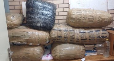 Eight large packages wrapped in black and brown tape are stacked against a brick wall. A map is partially visible above, with a plastic bottle nearby.