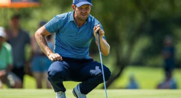 Golfer in a blue polka-dot shirt and cap crouches on a lush green golf course, focusing intently on the ball. The background is blurred with trees.