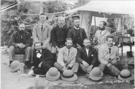 A black and white photo of nine men in suits and ties, sitting outdoors under a canvas tent. They appear formal and serious. Hats rest on the ground in front.