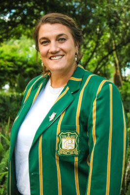 A smiling woman stands outdoors, wearing a green and yellow striped blazer with an emblem. Lush greenery forms the background, creating a cheerful, vibrant mood.