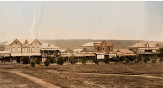 A sepia-toned historic town scene depicting a row of old buildings with distinct architectural designs. Small trees line the street, bordered by dry, grassy land.