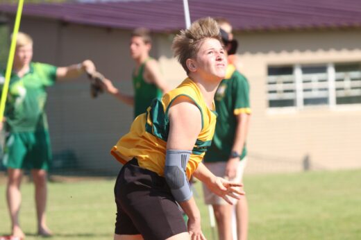 A young athlete in a yellow jersey, wearing an arm brace, focuses intensely while preparing to throw a shot put. Teammates and a building are blurred in the background.
