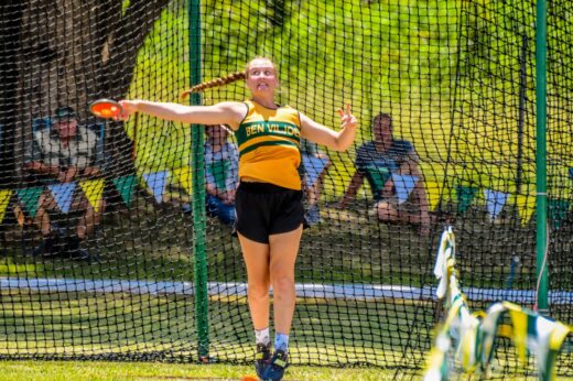 Athlete in a yellow and black uniform prepares to throw a discus in a netted field. Intense focus and sunny weather create an energetic atmosphere.