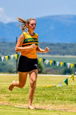 A focused young runner in a yellow and green jersey competes barefoot on a grassy track, set against a backdrop of mountains and blue sky.