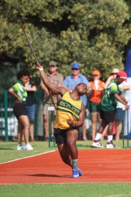Athlete in a yellow and green uniform throws a javelin on a red track, surrounded by spectators. The scene conveys focus and athleticism.