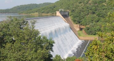 A large dam with water cascading down its smooth concrete surface, surrounded by lush green hills and dense foliage under a clear blue sky.