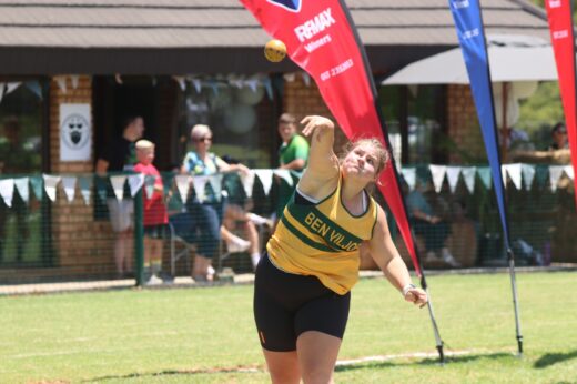 A female athlete in a green and yellow jersey throws a shot put with intense focus. Onlookers, banners, and a building are visible in the sunny background.