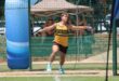 A male athlete in a yellow and green outfit prepares to throw a discus in a netted area, focused and poised, under sunny skies at a sports field.