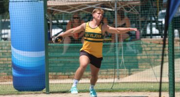 A male athlete in a yellow and green outfit prepares to throw a discus in a netted area, focused and poised, under sunny skies at a sports field.