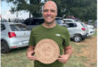 A smiling man in a green shirt holds a wooden plaque with a bicycle engraving and text reading "Mpumalanga Road Champs 2023 1st." Parked cars are in the background.