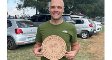 A smiling man in a green shirt holds a wooden plaque with a bicycle engraving and text reading "Mpumalanga Road Champs 2023 1st." Parked cars are in the background.