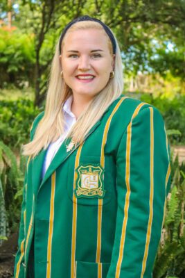 A smiling person in a green blazer with yellow stripes and an embroidered crest stands outdoors. The background is a lush, sunlit garden, conveying a bright and cheerful mood.