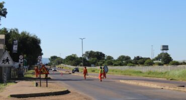 Construction workers in orange uniforms set up road signs on a sunny day. A car approaches in the distance. Trees and a water tower are visible.