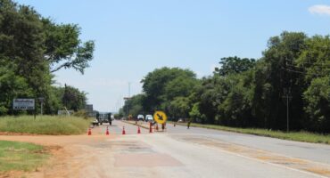 A road with a construction sign indicating left at a work zone. Orange cones line the area, with green trees and bushes on each side under a clear blue sky.