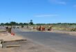 Road construction with workers in orange safety gear on a sunny day. Traffic cones line the road, with a clear blue sky and distant greenery.