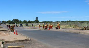 Road construction with workers in orange safety gear on a sunny day. Traffic cones line the road, with a clear blue sky and distant greenery.