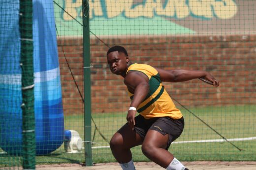 Athlete in a yellow and green jersey prepares to throw a discus. The scene is outdoors on a sunny day with a net and blurred background. Focused expression.