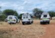 Three police vans are parked on a dirt road in a rural area with dry grass and scattered trees under a cloudy sky, conveying a serious, official tone.