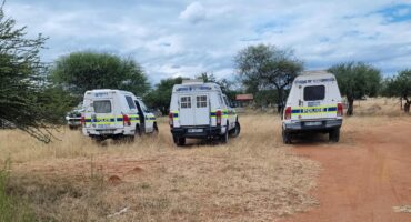 Three police vans are parked on a dirt road in a rural area with dry grass and scattered trees under a cloudy sky, conveying a serious, official tone.