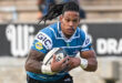 Rugby player in a blue and white jersey fiercely runs holding a rugby ball. His focused expression and dreadlocks convey strength and determination.