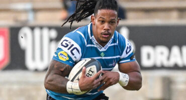 Rugby player in a blue and white jersey fiercely runs holding a rugby ball. His focused expression and dreadlocks convey strength and determination.