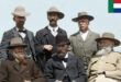 A group of six men in historical attire pose outdoors. Four are seated, two stand behind. They wear hats and suits, creating a formal and serious tone.