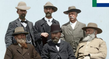 A group of six men in historical attire pose outdoors. Four are seated, two stand behind. They wear hats and suits, creating a formal and serious tone.