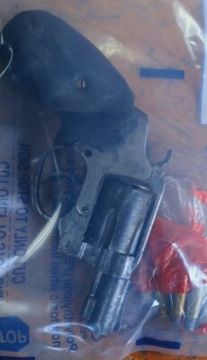 A black revolver with a textured grip and several bullets are sealed in a transparent evidence bag. The scene has a serious, forensic tone.