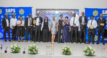 A group of individuals, dressed formally, stand on a stage holding awards. Two "SAPS" banners are in the background. The mood is celebratory.