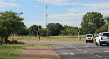 A sunny intersection with traffic lights, bordered by lush trees and a grassy field. Two vehicles are waiting. The scene is calm and open-air.