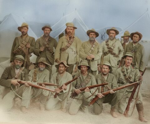 A sepia-tone photo shows 14 soldiers in rugged uniforms and hats, holding rifles. They stand and kneel in front of white tents, conveying a historical military scene.