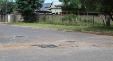A residential street with a few visible potholes. In the background, there are trees and a house surrounded by a wall and a fence. The mood is calm.