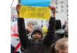 Child holding "#StandWithUkraine" sign at a protest. He wears a gray beanie and jacket. Other protestors with signs are visible in the background.