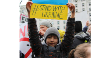 Child holding "#StandWithUkraine" sign at a protest. He wears a gray beanie and jacket. Other protestors with signs are visible in the background.