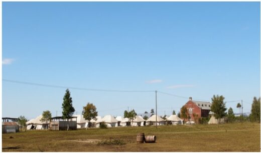 A row of white tents and a red brick building sit under a clear blue sky, surrounded by trees and an open grassy field. Two barrels are in the foreground.