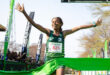 Runner in a green uniform, bib number 825, jubilantly crosses a bright green finish line. Banners and cheering crowd in the background.