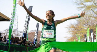 Runner in a green uniform, bib number 825, jubilantly crosses a bright green finish line. Banners and cheering crowd in the background.