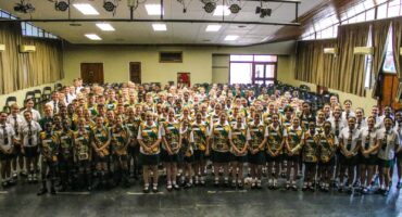 A large group of students in school uniforms stand closely together in a school hall. The mood is cheerful and lively, with smiling faces and bright lighting.