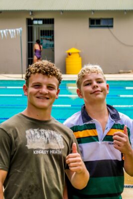 Two boys stand smiling in front of a swimming pool, giving thumbs up. One wears a striped shirt and the other a t-shirt. A relaxed and cheerful scene.